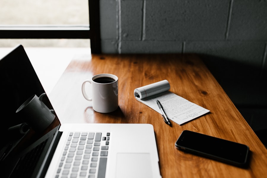 A laptop, coffee and notebook on a wooden desk by a window